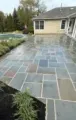 Stone patio beside a backyard pool and beige house, with garden beds and overcast sky.