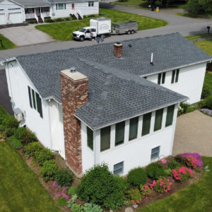 Aerial view of a white house with a brick chimney, gray roof, and pink flower bed on a curved driveway.