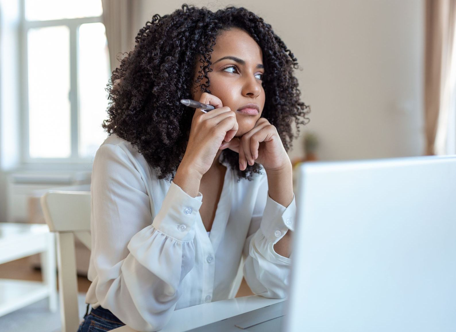 An African American woman sits in front of a laptop, she looks forlornly out the window, she does not yet know the reasons to hire a life coach