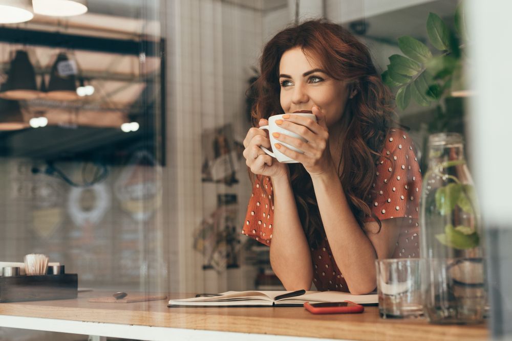 A woman is sitting at a table drinking a cup of coffee and looking out the window.