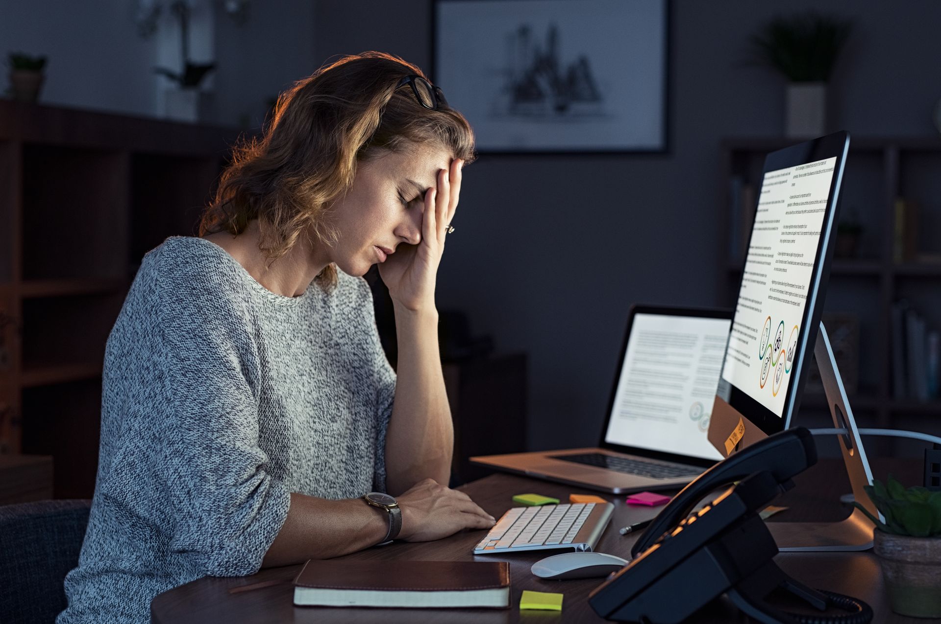 A middle aged woman sits at her desk in the dark, rubbing her temples she is suffering from the 5 stages of burnout