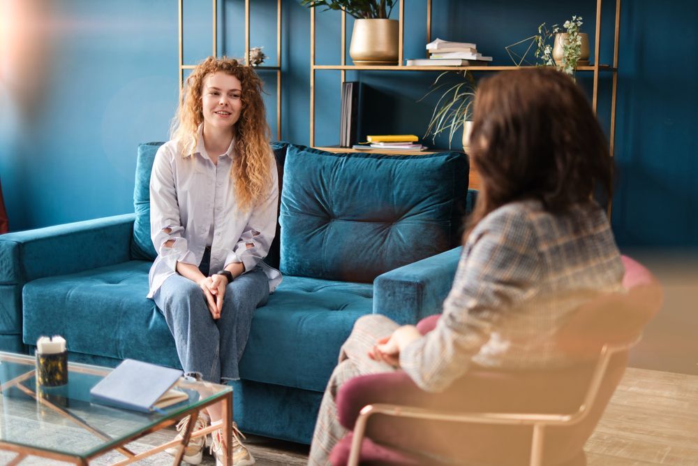 A woman is sitting on a couch talking to another woman.