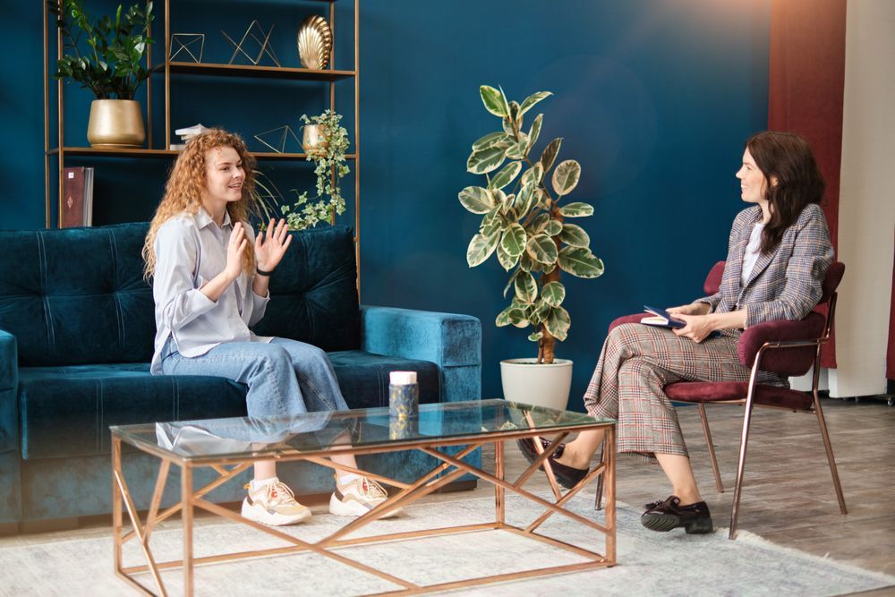 Two women are sitting on a couch and talking to each other in a living room.