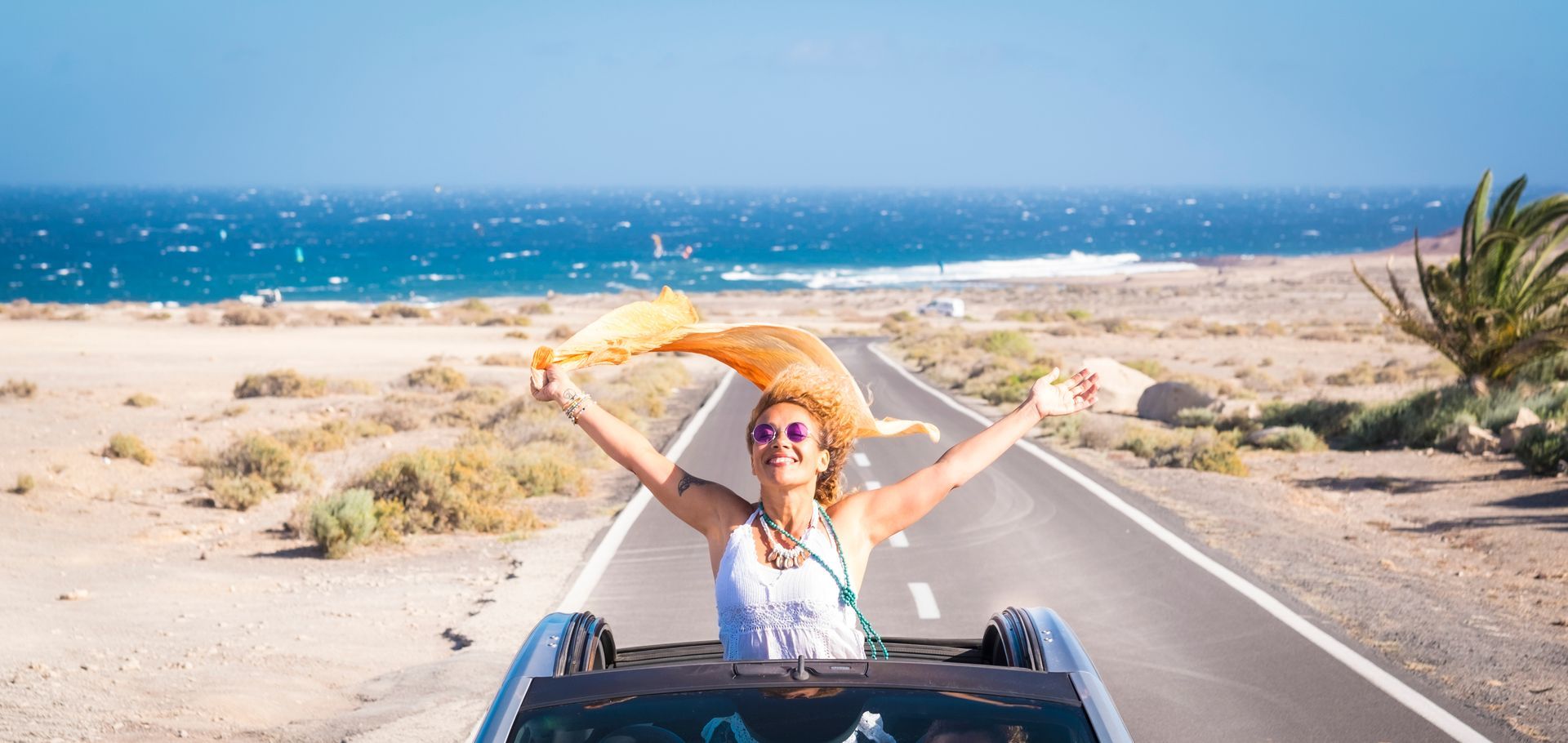 A woman with curly hair stands up out of a convertible holding an orange cloth, she contemplates the difference between self-confidence vs self-esteem