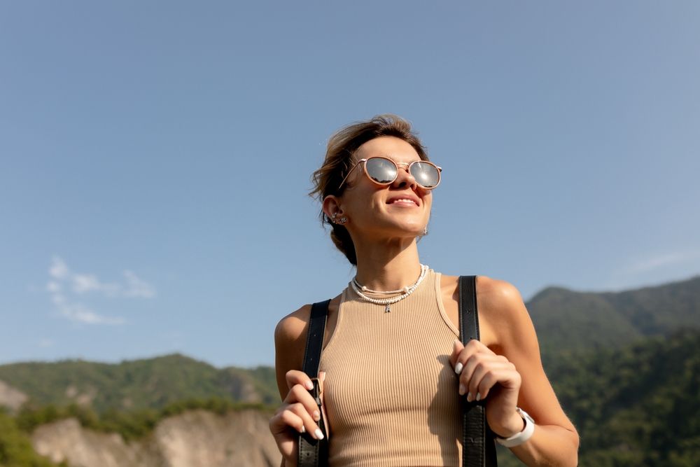 A woman wearing sunglasses and a backpack is standing on top of a mountain.