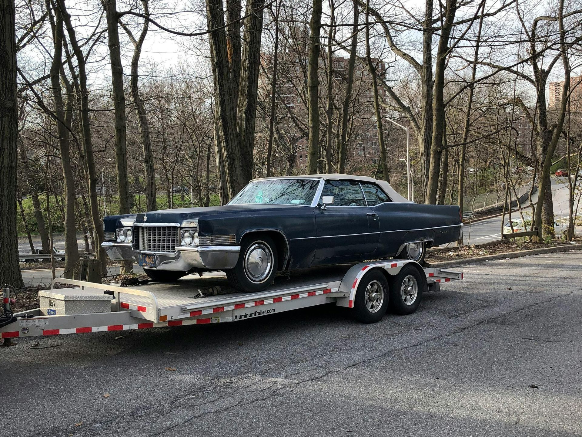 A vintage blue Cadillac convertible sits on a trailer in a wooded area. The car has a white top and chrome details.