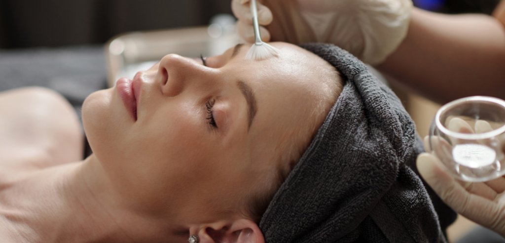 A person receiving a facial treatment at a spa. A technician applies a product to their forehead with a small brush.