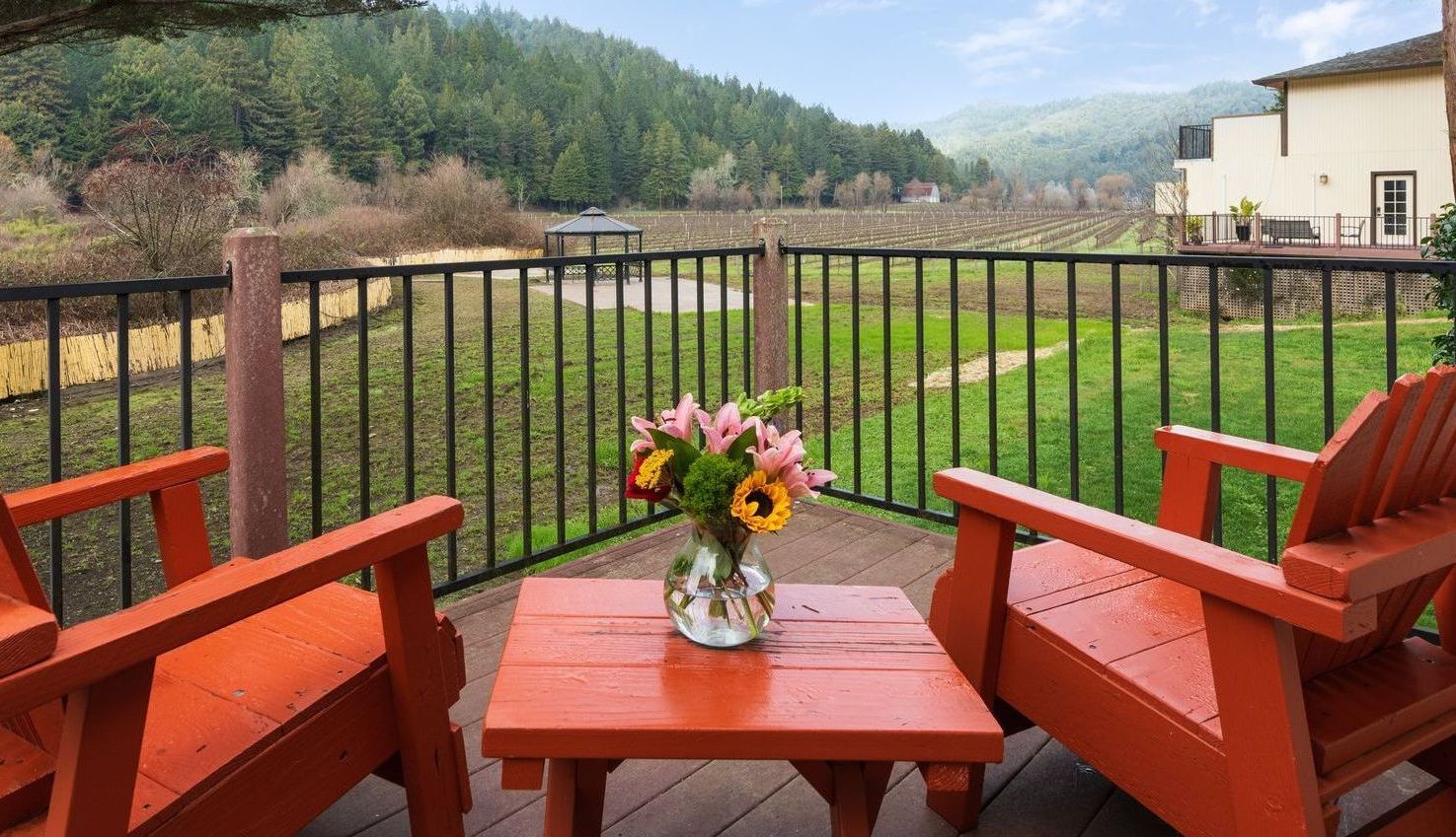 Two red Adirondack chairs and a small table with flowers on a deck overlooking a vineyard and rolling hills.