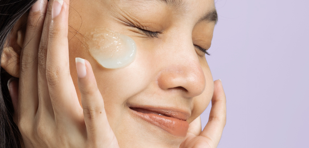 A person smiles with closed eyes and a skincare product on her cheek.