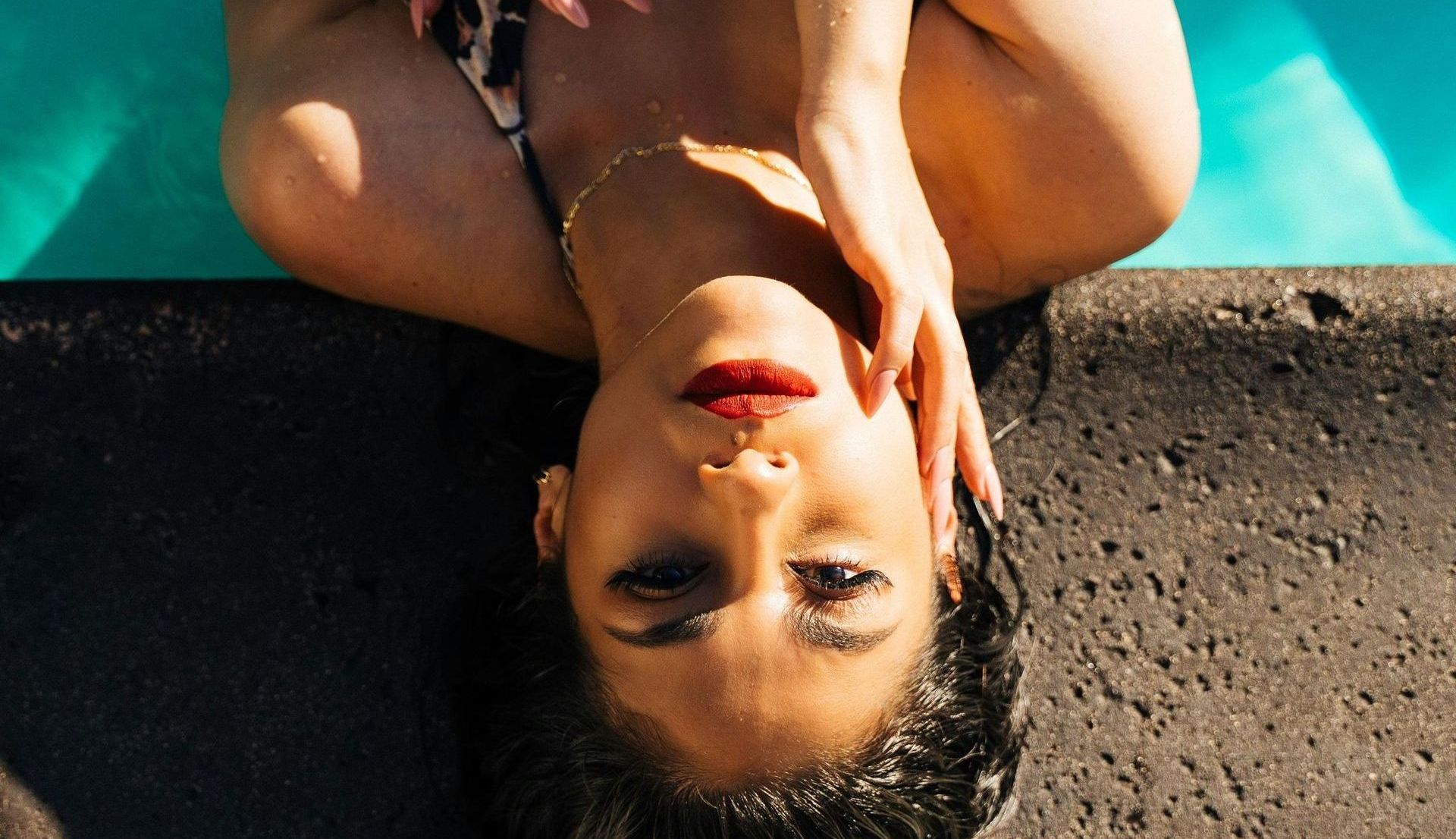 Woman relaxes in pool with head back on cement, looking at camera