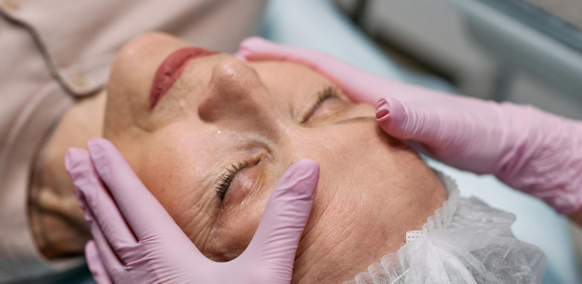 A person with closed eyes having their face gently touched by two hands wearing pink gloves, likely for a beauty treatment.