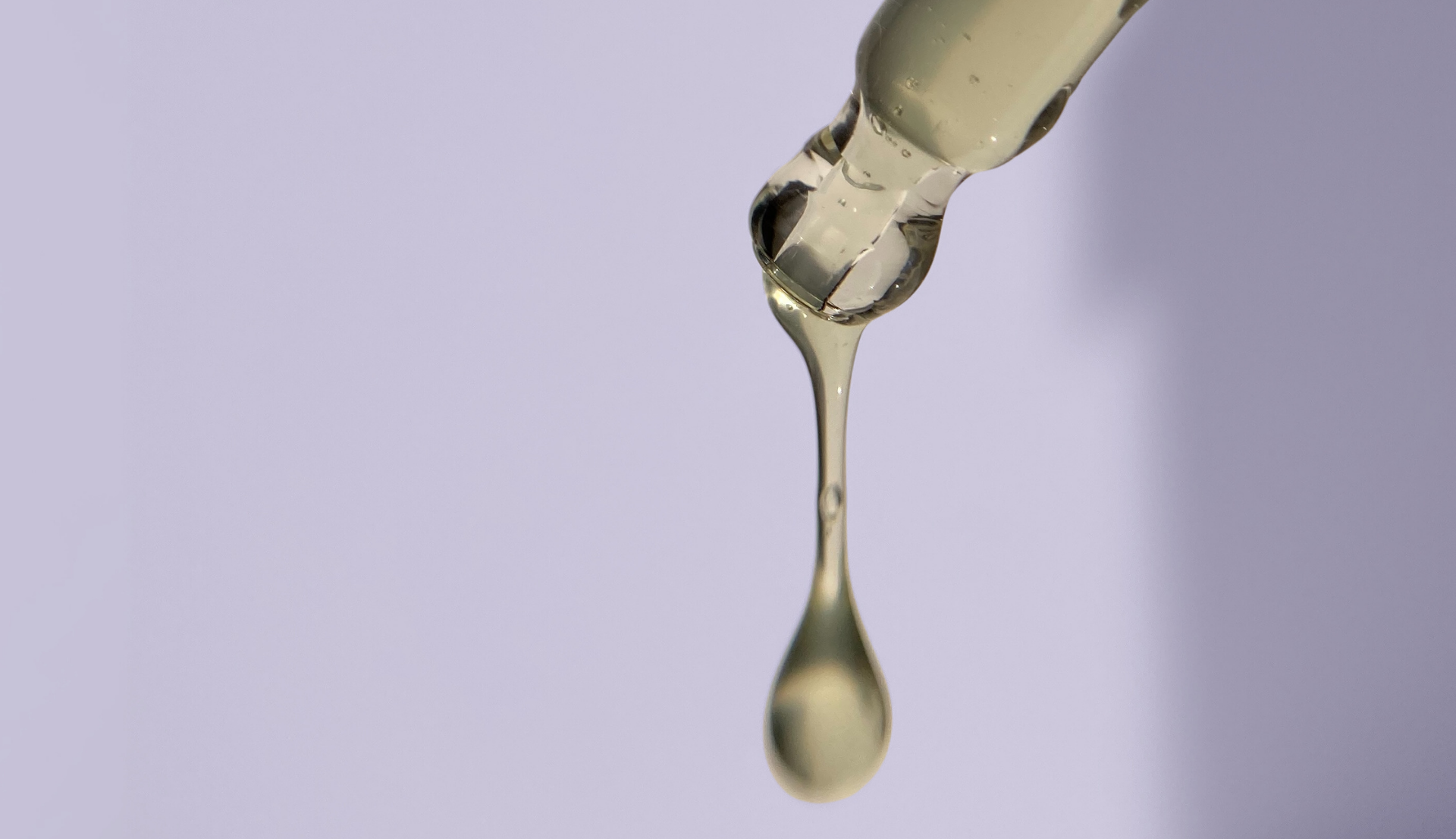 A dropper dispensing a single, viscous, yellow liquid droplet against a light purple background.