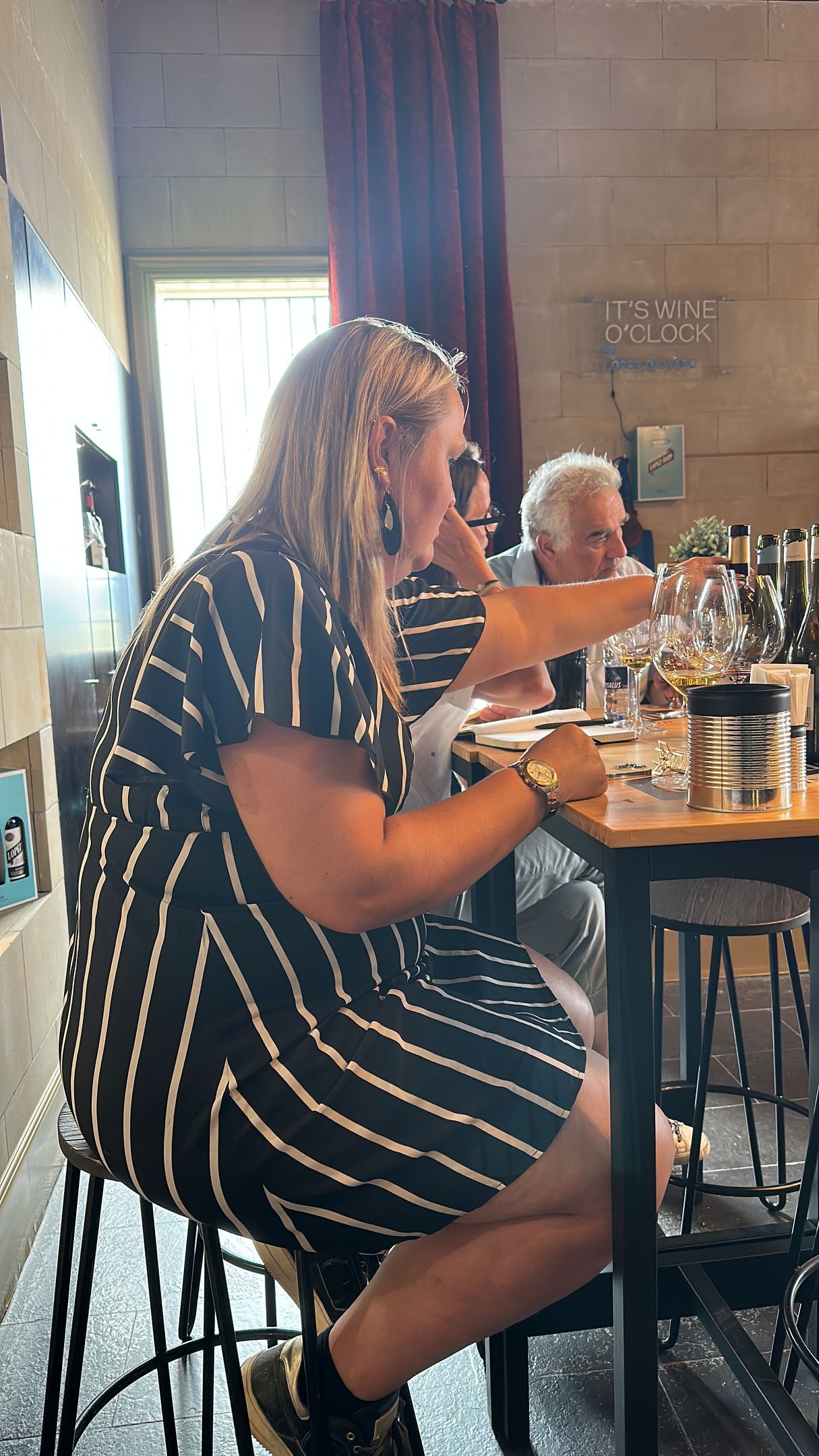 Vrouw in gestreepte jurk aan tafel, gebarend, met oudere man. Binnen, zonlicht.