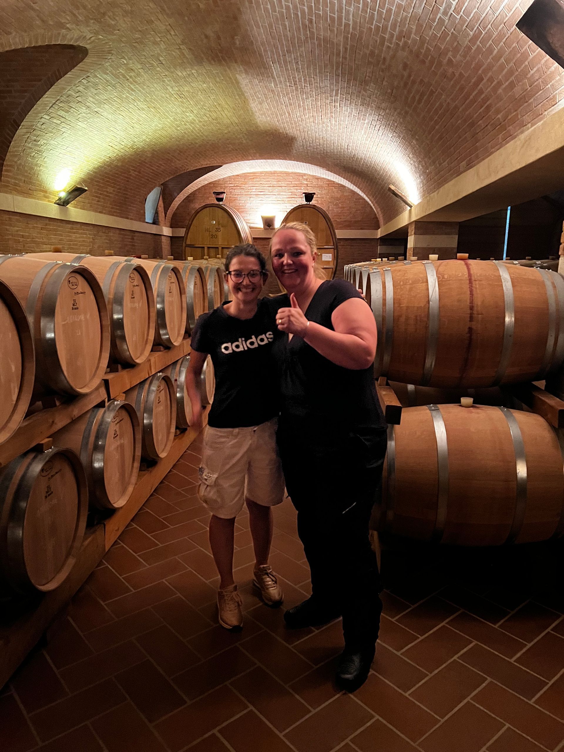 Twee vrouwen staan ​​in een wijnkelder bij houten vaten; een van hen steekt haar duim op.