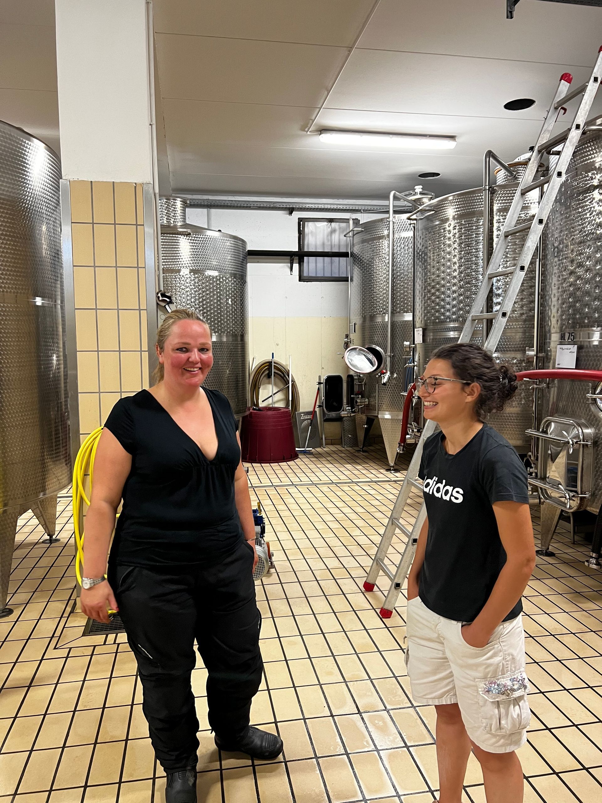 Twee lachende vrouwen in een wijnkelder met grote stalen tanks.