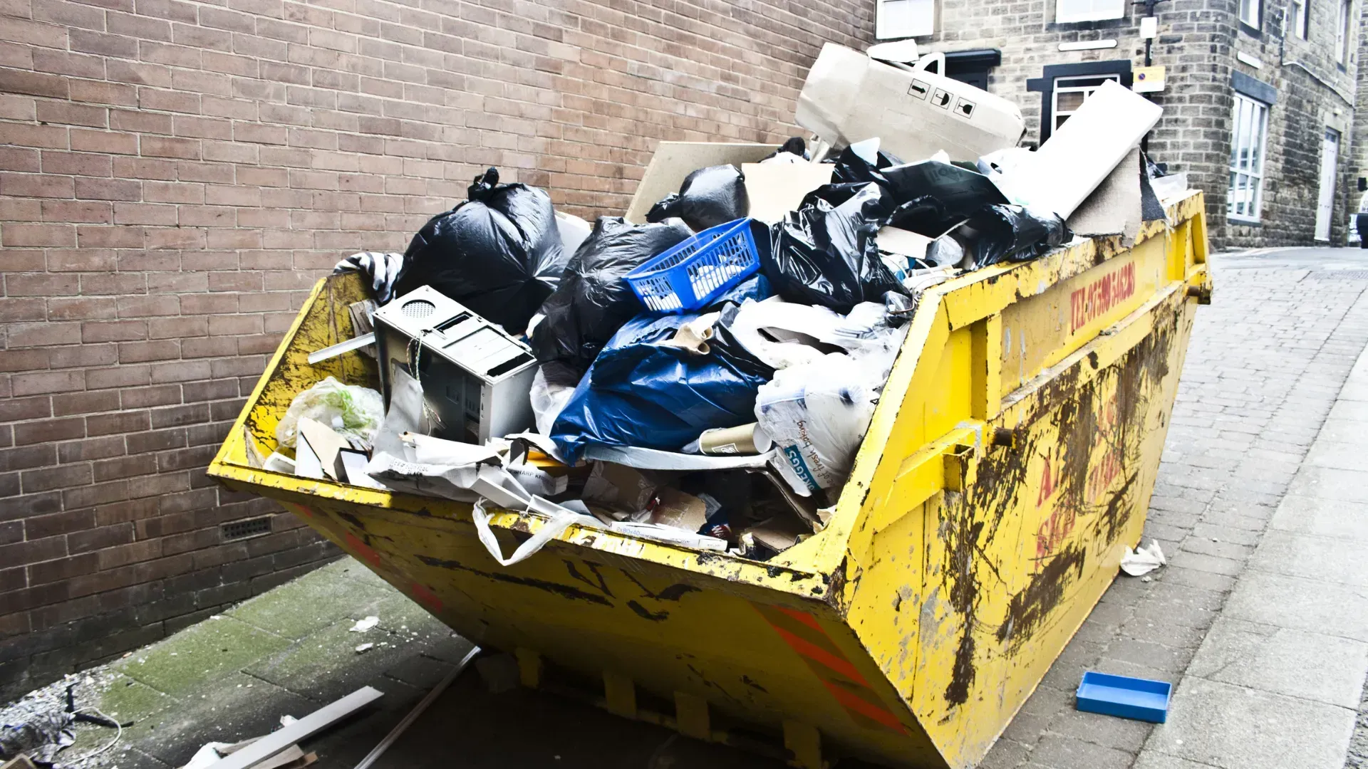 A yellow skip overflowing with garbage bags, cardboard, and various debris, located on a sidewalk next to a brick wall.