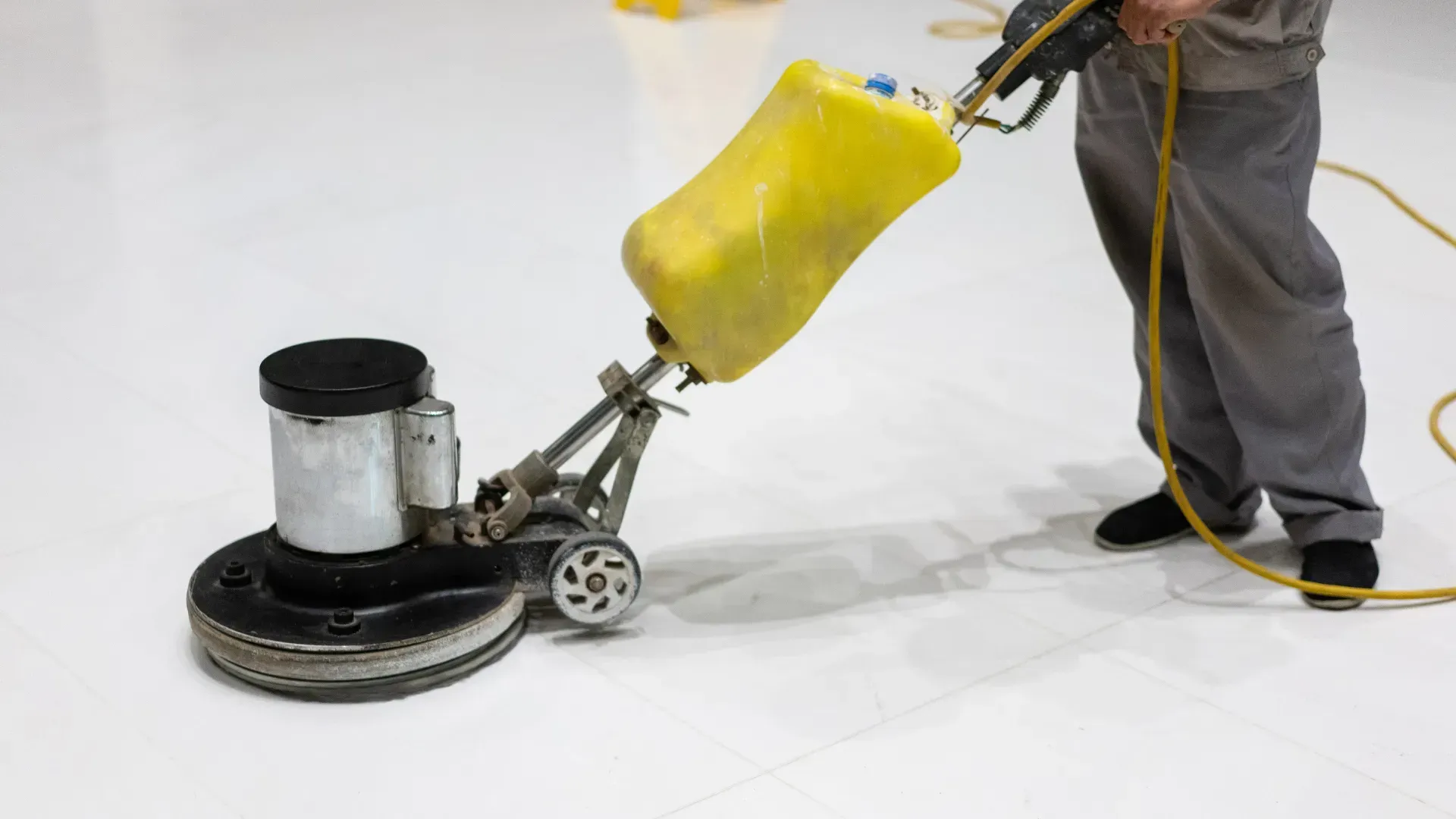 Person using a yellow and black floor cleaning machine on a white floor. The machine is in operation.