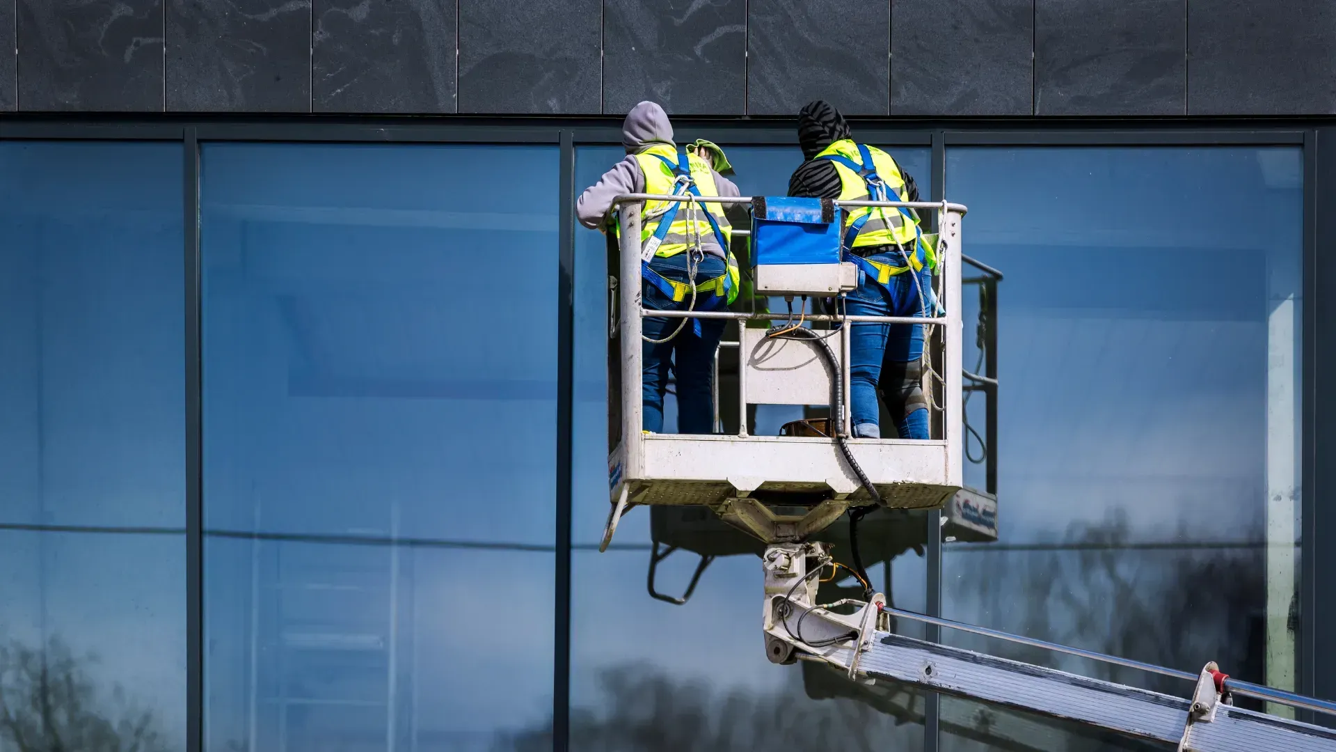 Two workers in safety vests on a lift platform are inspecting a glass building facade. The lift is extended upwards.