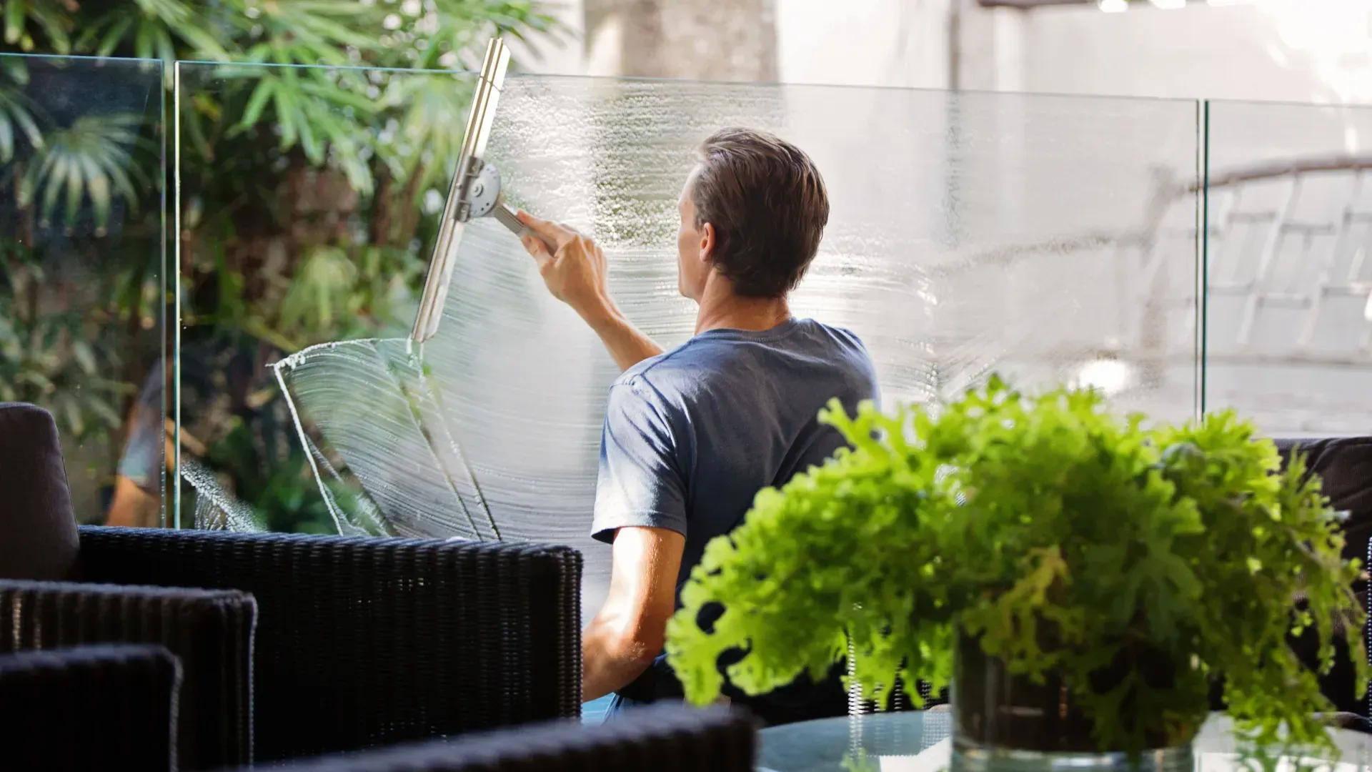 Man using a squeegee to clean a glass panel outdoors. Green plants and wicker furniture are in the foreground.