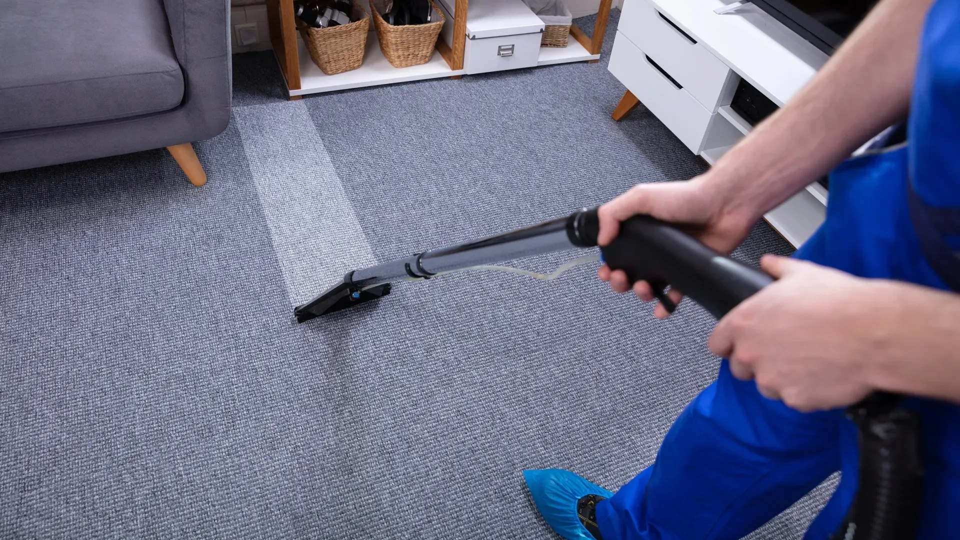Person in blue cleaning a gray carpet with a vacuum, leaving a clean strip behind. Interior setting.