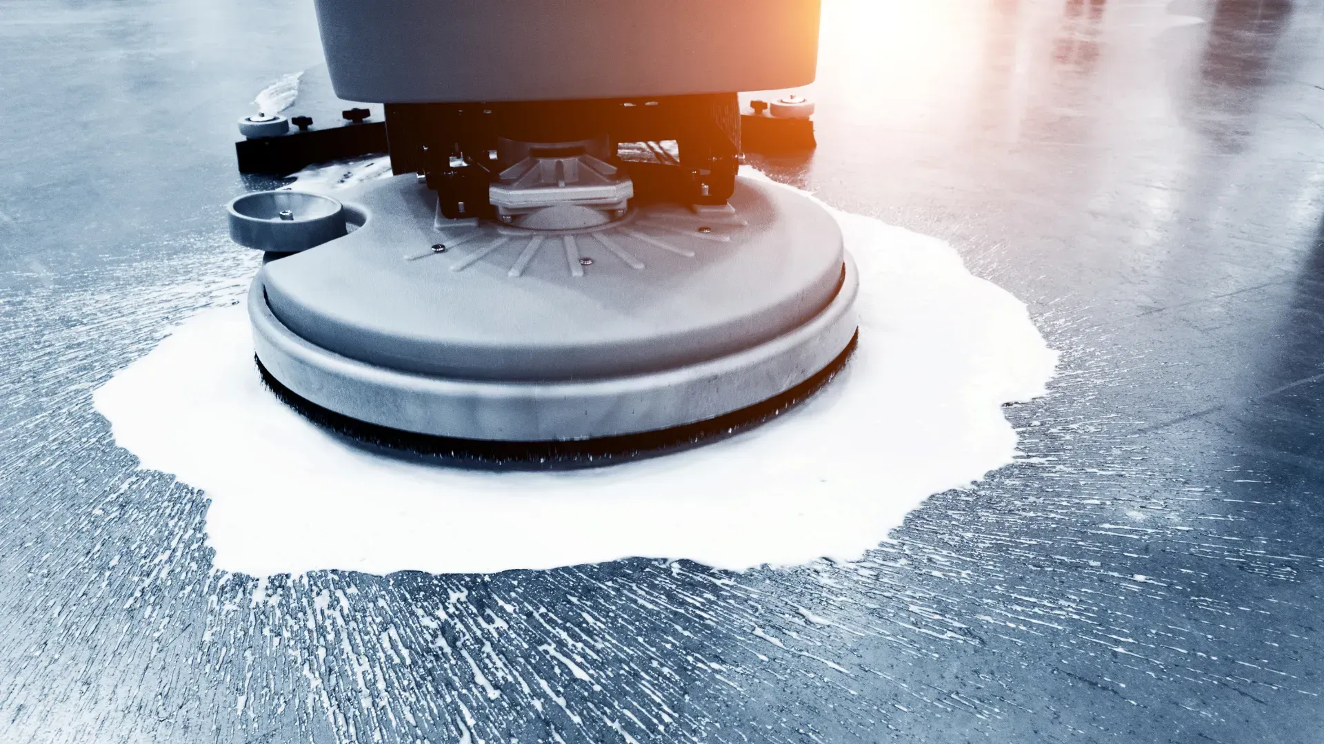 A floor cleaning machine in action, spraying cleaning solution. Bright light and bluish-gray floor.