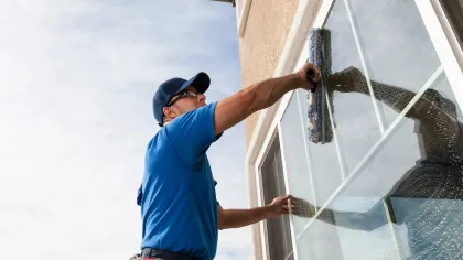 Man in blue shirt and cap cleaning a window with a squeegee, against a building exterior and cloudy sky.
