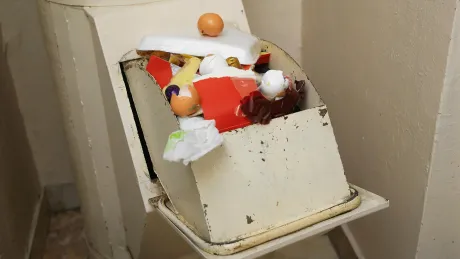 Overflowing trash can in a light-colored enclosure, filled with food waste, a carton, and a single egg on top.
