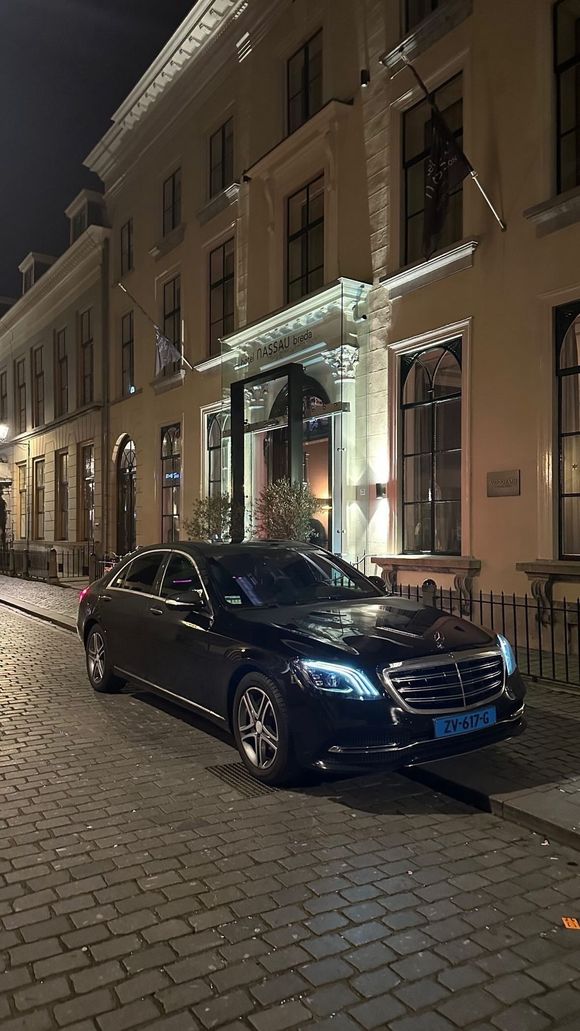 Een elegante zwarte Mercedes-Benz staat geparkeerd op een straat met kinderkopjes voor een gebouw dat 's nachts verlicht is met lichtsnoeren.