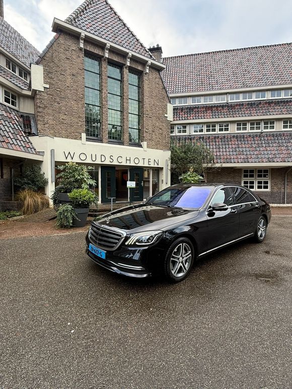 Een zwarte luxe sedan staat geparkeerd op een grindpad voor het Wolfheze hotel, dat een bakstenen gevel heeft.