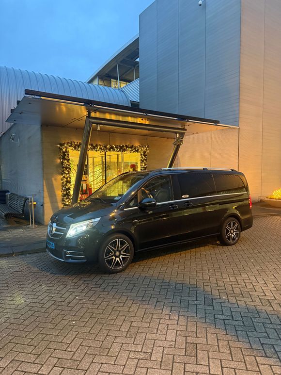 Een donkergrijze Mercedes-Benz V-Klasse personenwagen staat geparkeerd op een verharde oprit voor een gebouw met verlichting in de schemering.