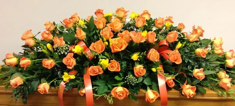 A large arrangement of orange roses and yellow daisies on a coffin.