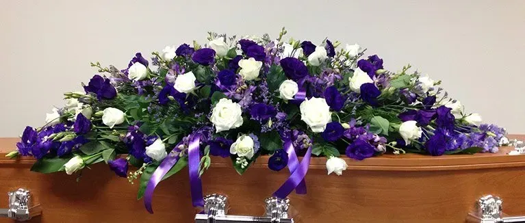 A coffin is decorated with purple and white flowers and ribbons.