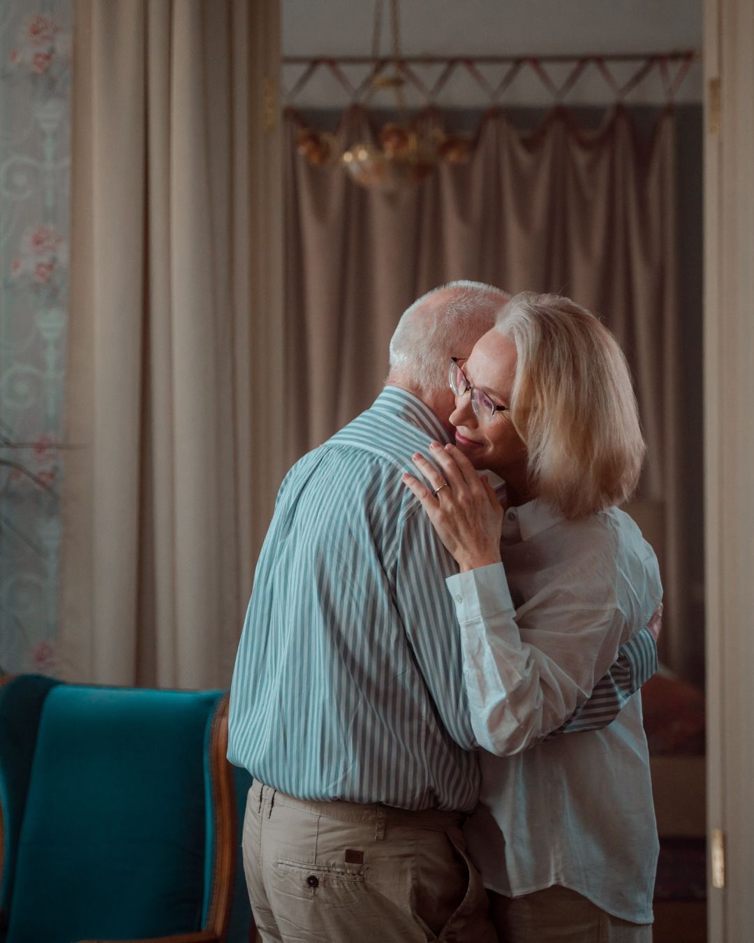 An elderly couple is hugging each other in a living room.