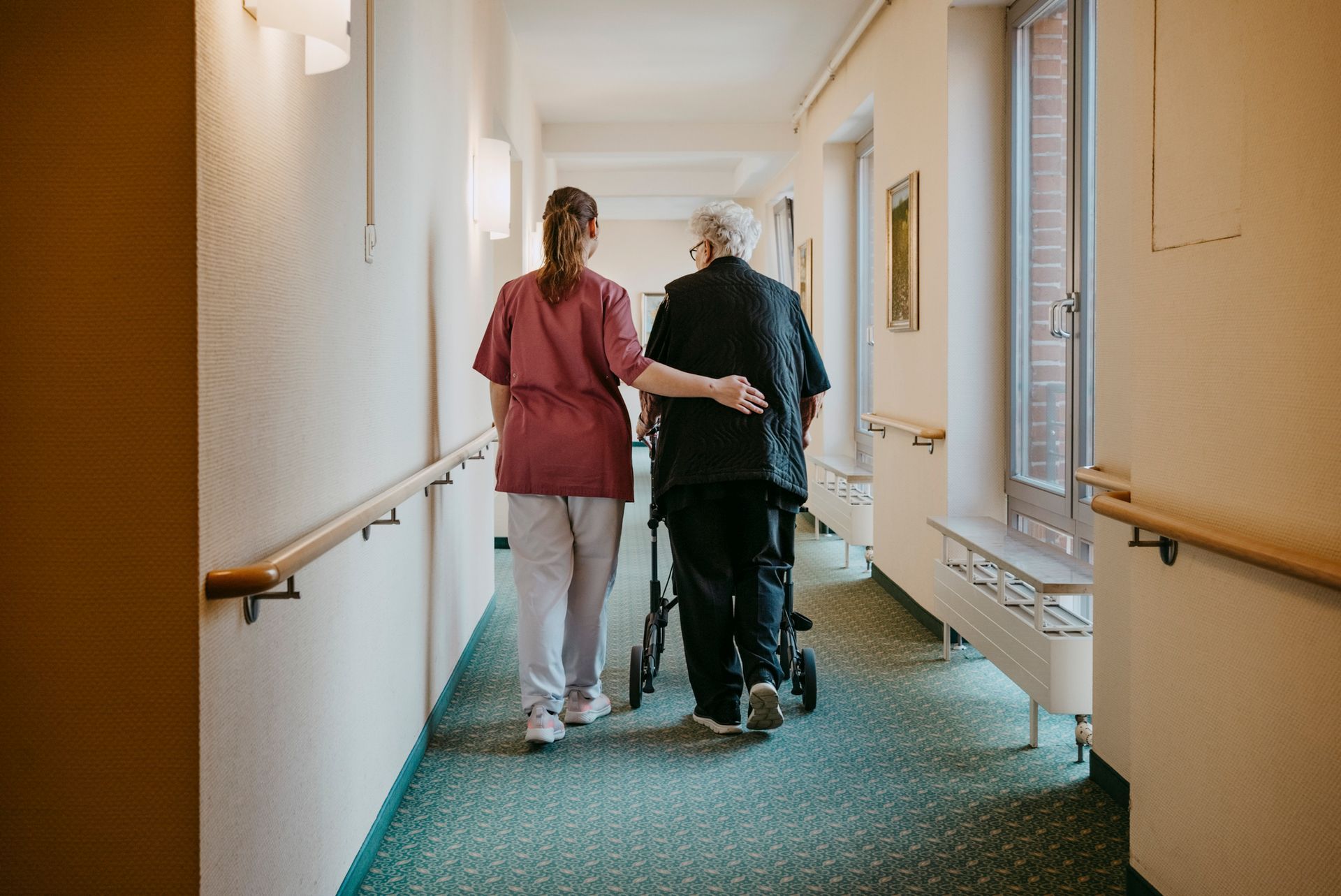 A nurse is helping an elderly man with a walker down a hospital hallway.