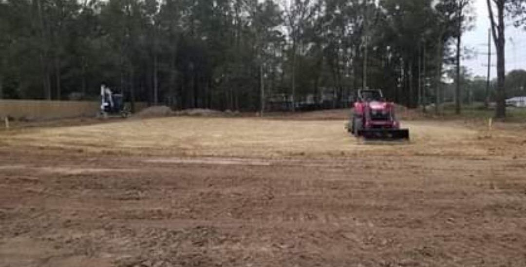 A red tractor is plowing a dirt field with trees in the background.