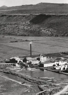 Factory with tall smokestack, pond, and surrounding landscape.