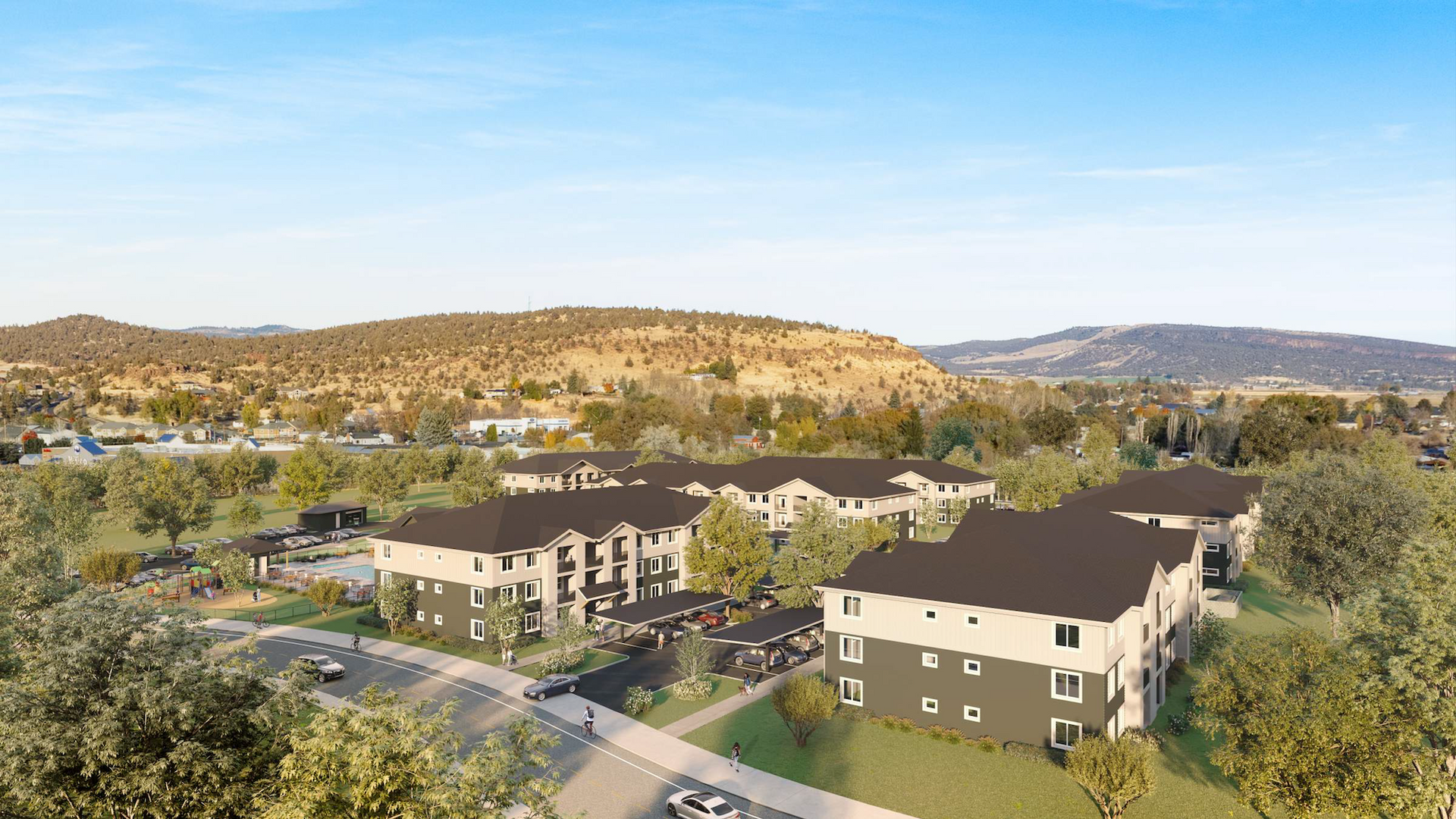 An aerial view of multiple apartment buildings with dark gray roofs, surrounded by trees and a hilly landscape.