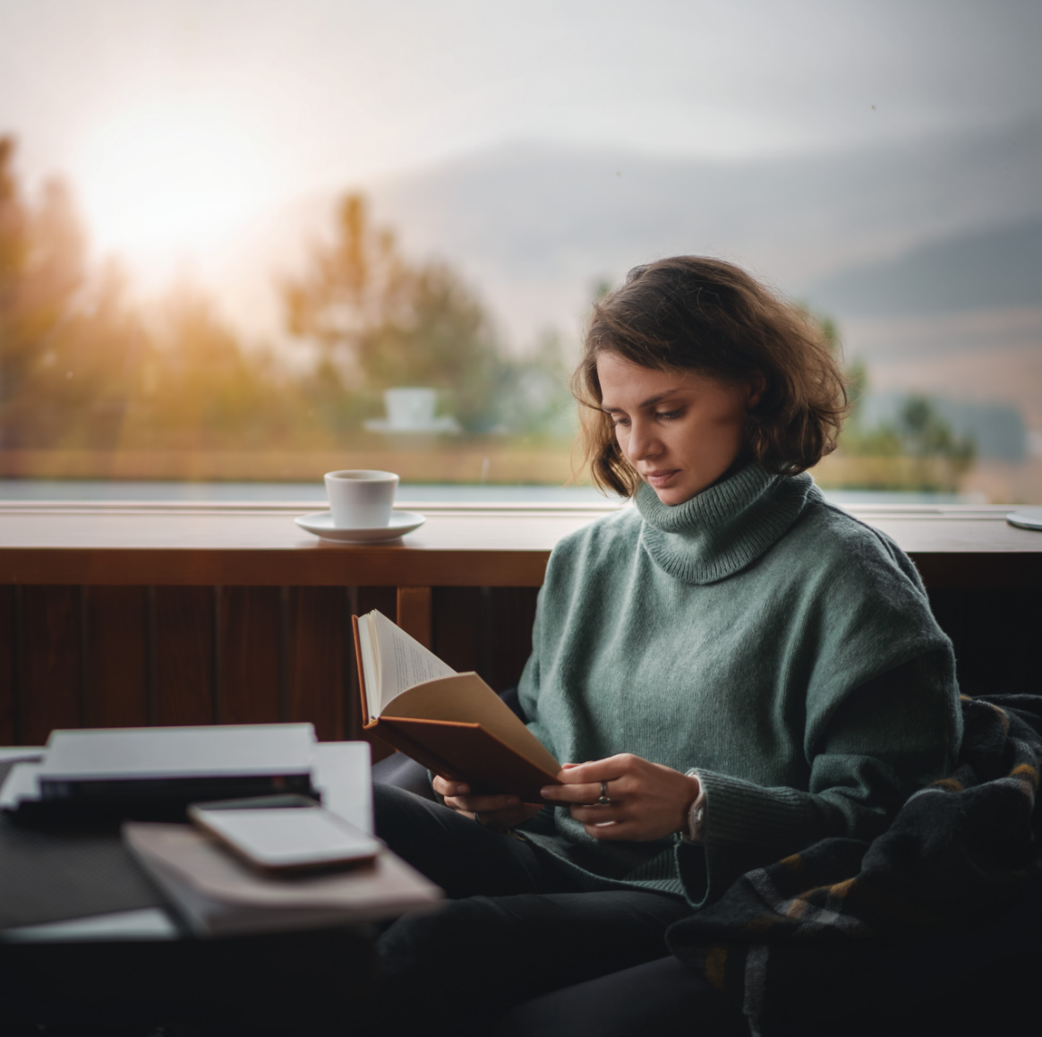Woman reading a book by a window, wearing a green sweater, with a cup of coffee nearby.