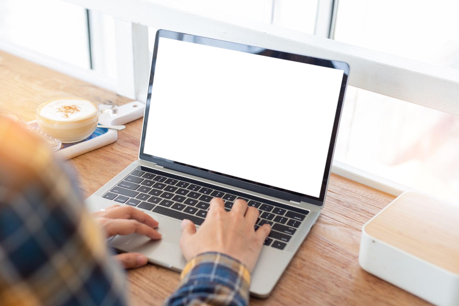 Person typing on a laptop with a blank screen at a wooden desk near a window. Cup of coffee nearby.