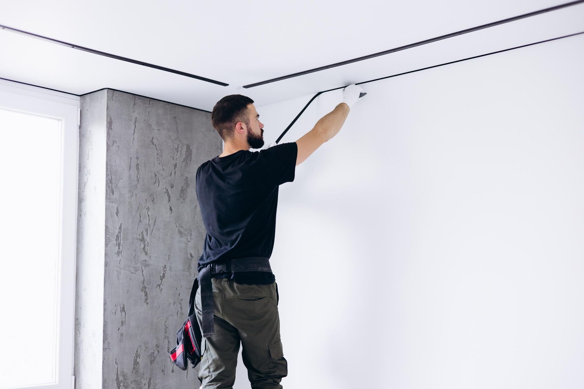 A person on a ladder installs black trim on a white wall in a room.