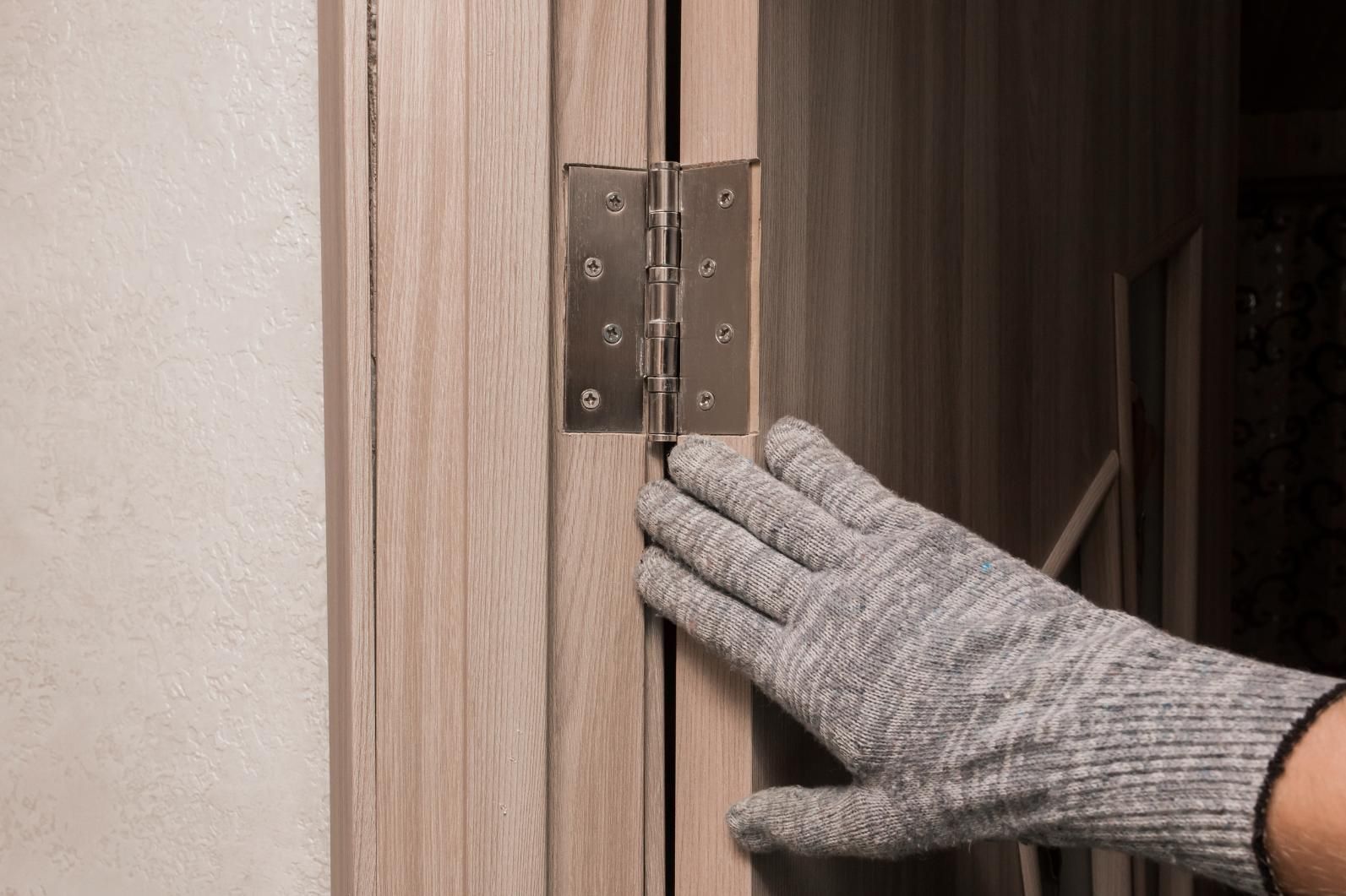 Gloved hand touching a door hinge on a wooden door frame, possibly checking for alignment or damage.