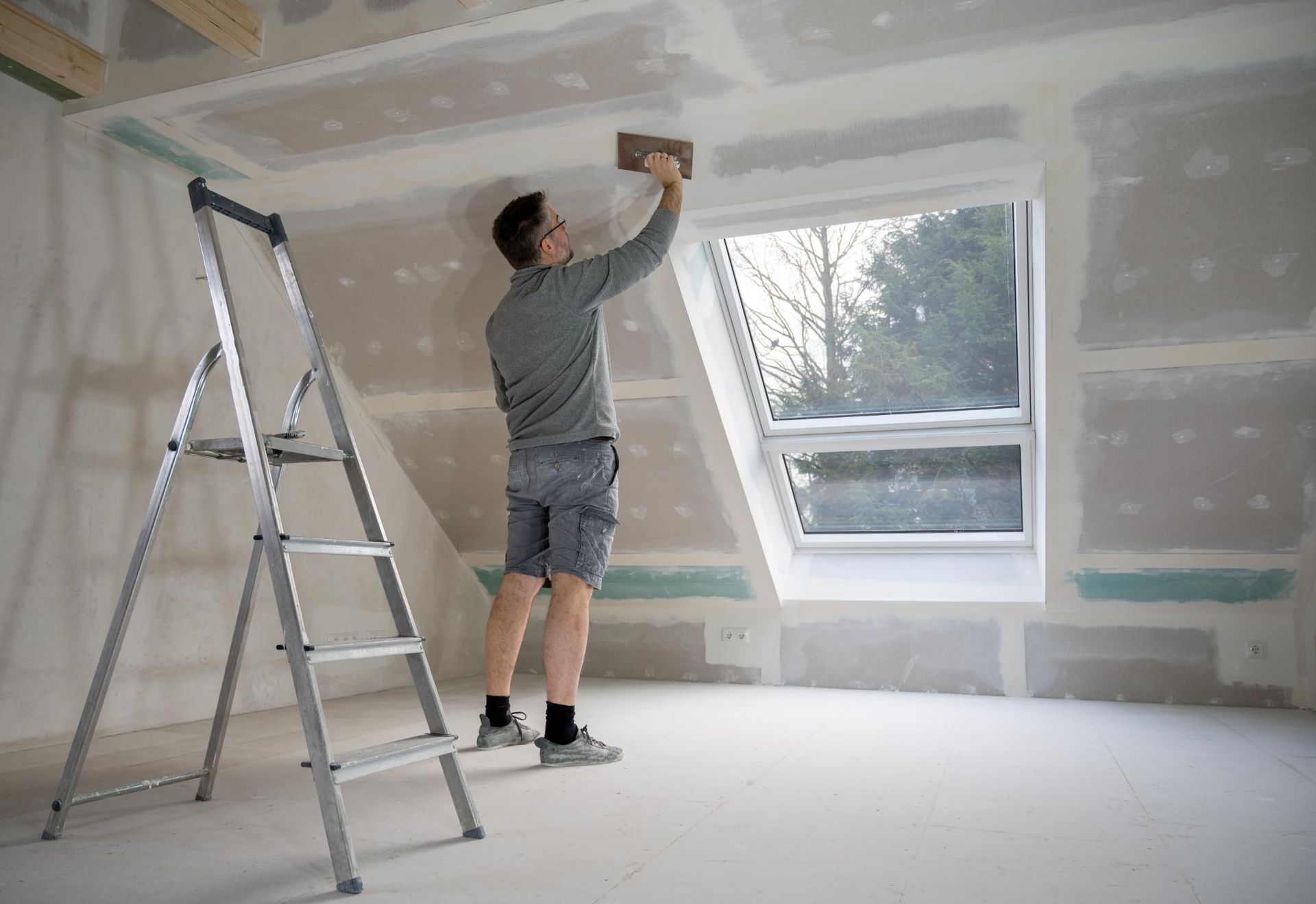 Person applying joint compound to drywall near a window, using a ladder in a room.