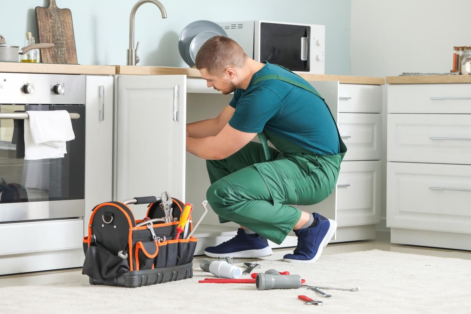 Plumber working under a kitchen sink, tools at his feet. He wears green overalls and a blue shirt.
