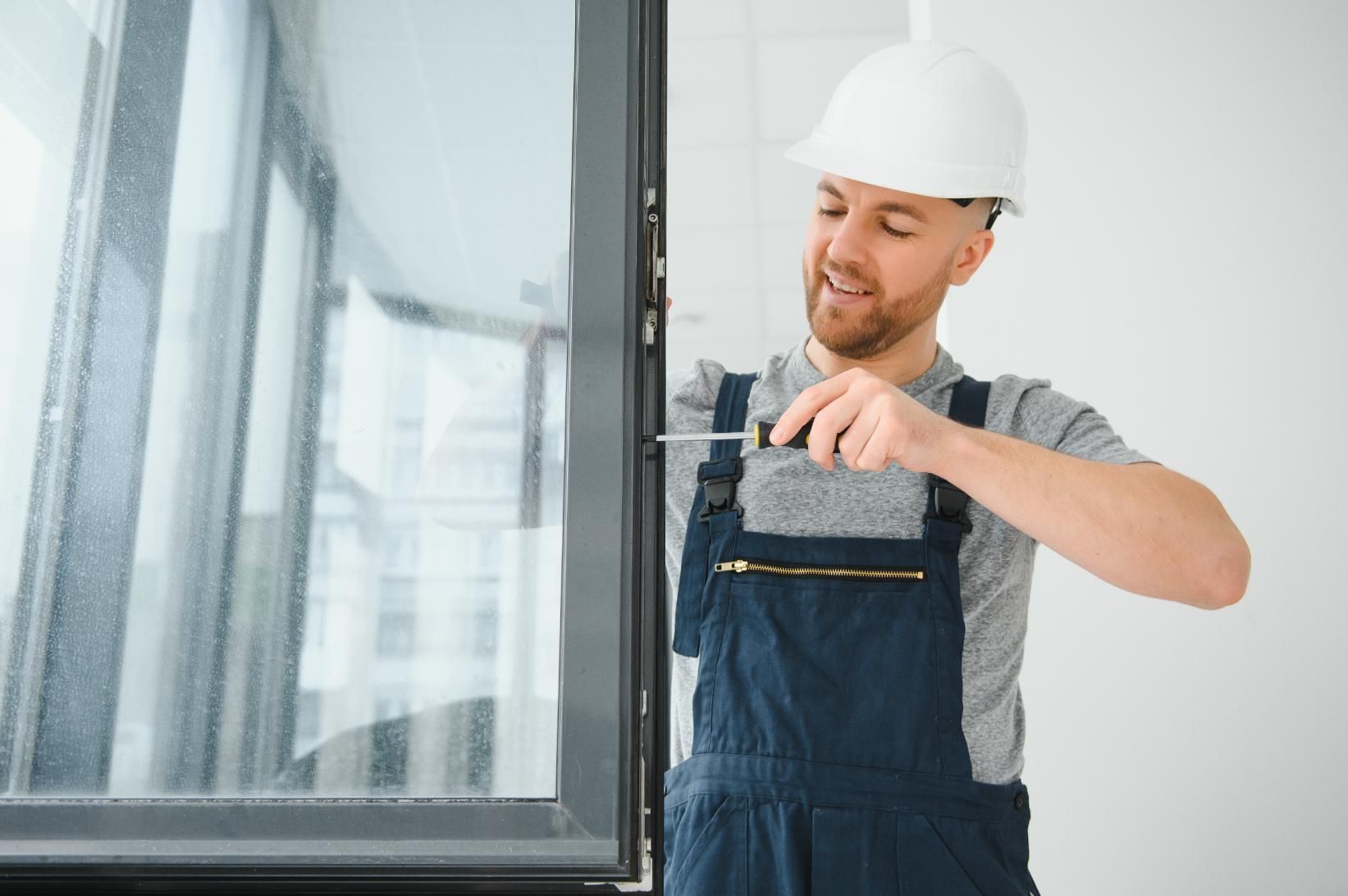 Man in work overalls and hard hat using a screwdriver on a window frame.