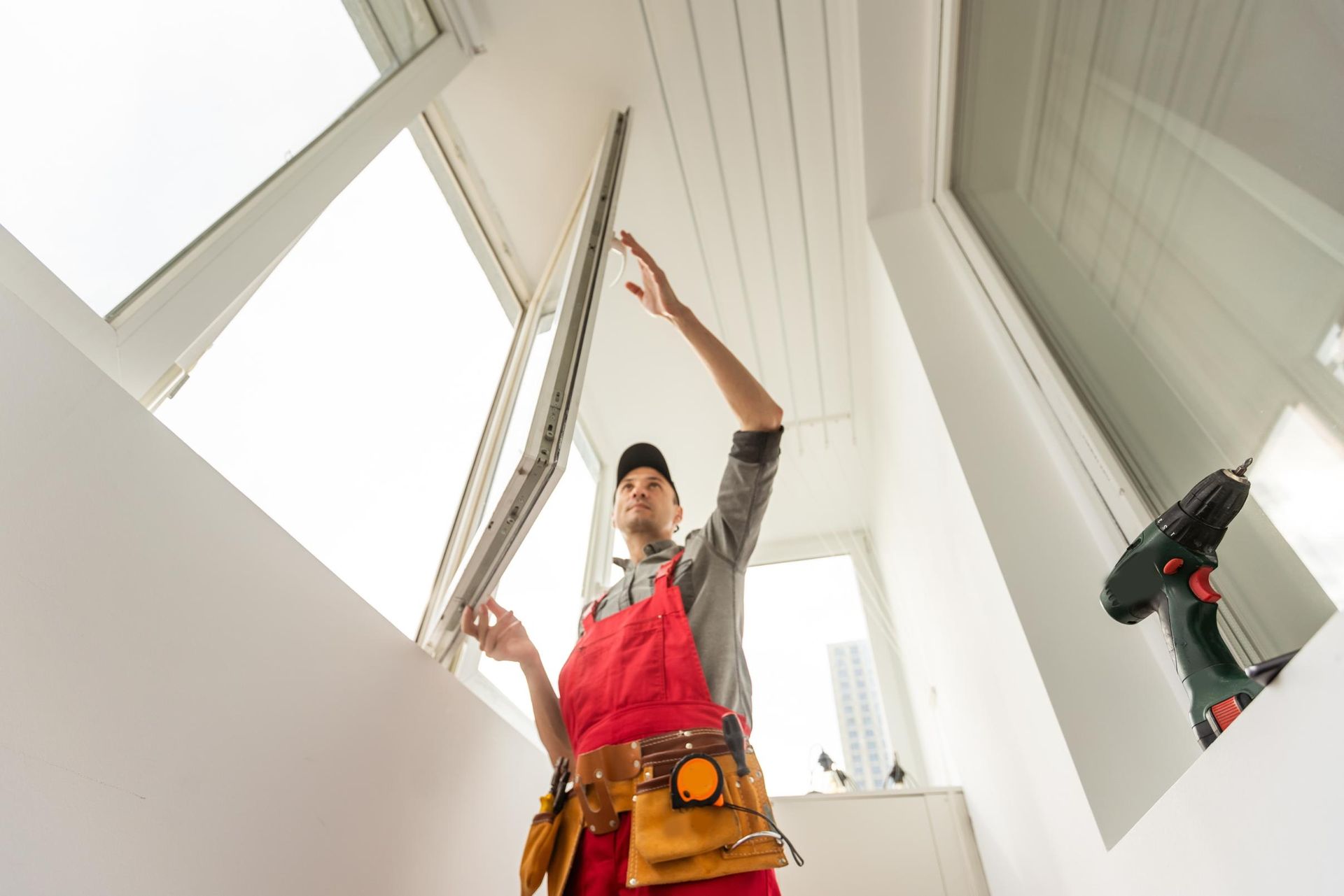 A person in red overalls installs a window in a white room. A drill rests on the sill.