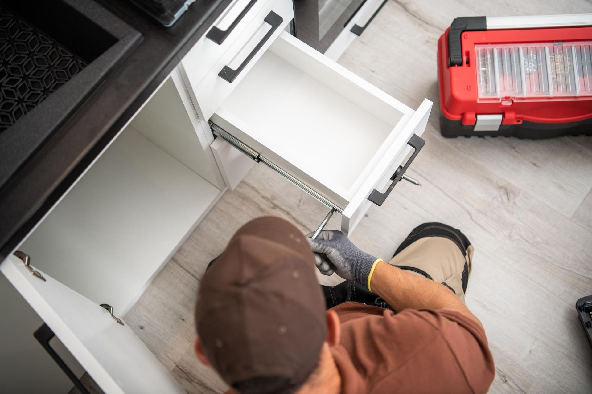 Person installing a drawer in a white kitchen cabinet, with a toolbox nearby.