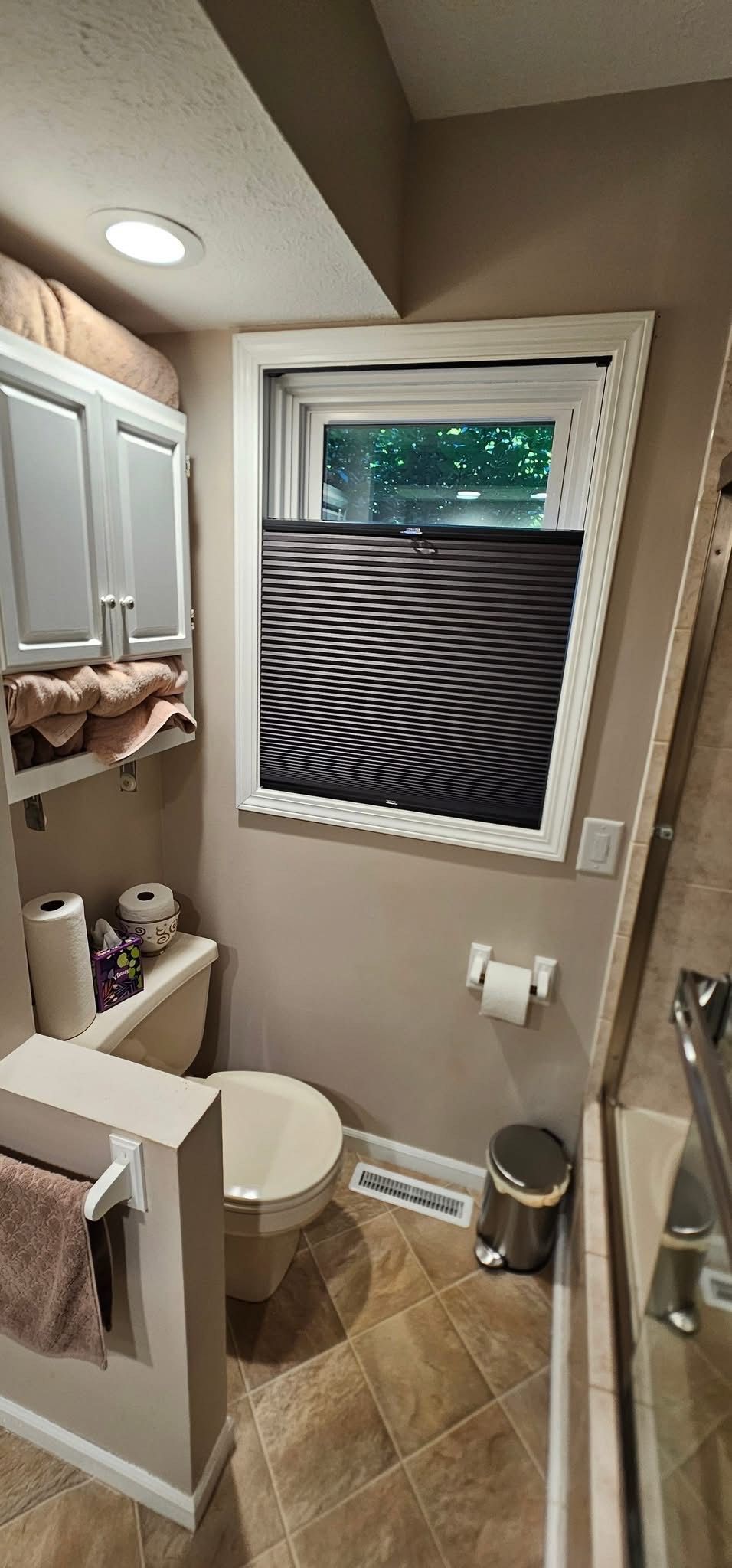Bathroom with toilet, window with patterned blind, and cabinet above.