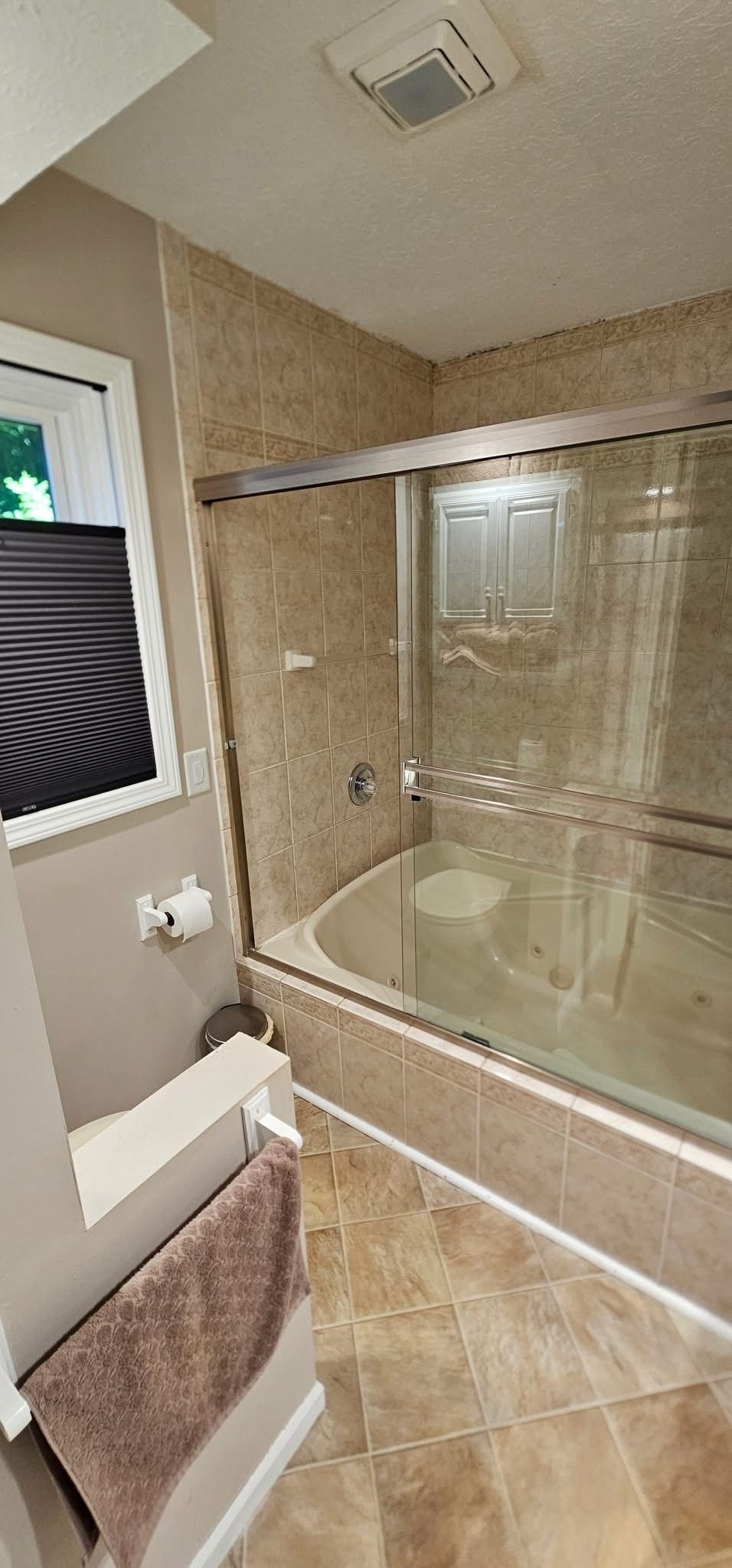 Bathroom with a tiled tub/shower, toilet, and window. Beige walls and floor. A towel hangs on the toilet.