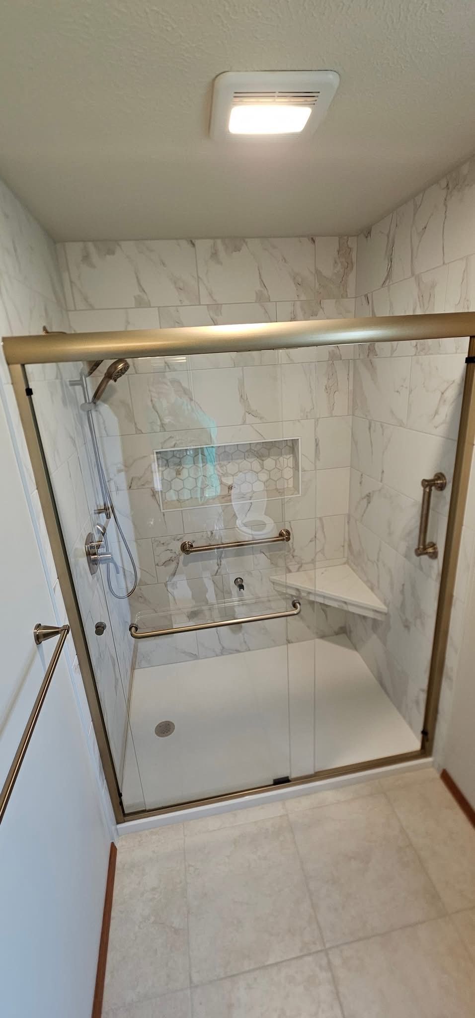 A shower with marble-look tiles, glass door, and brass hardware. Includes a built-in bench.