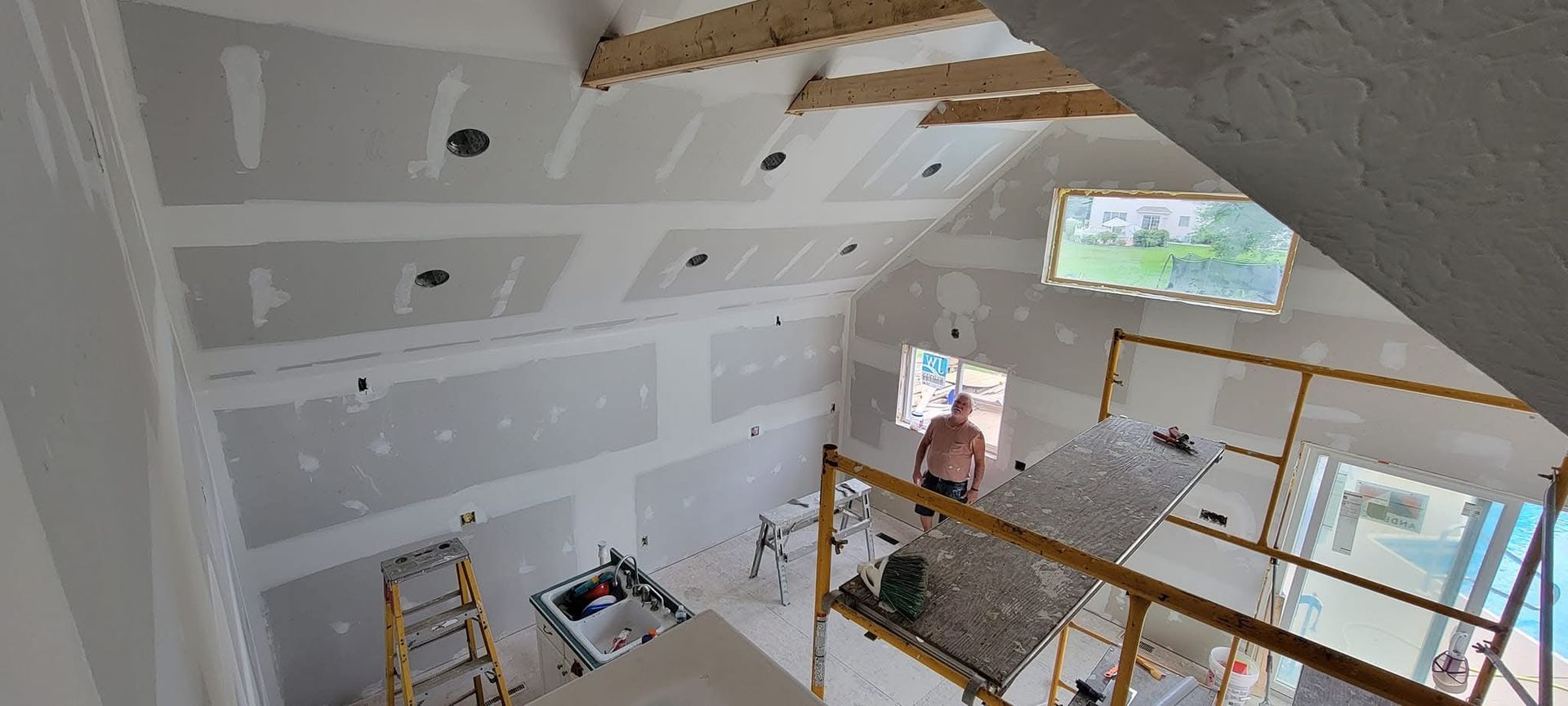 Construction site with drywall installed on walls and ceiling. A person stands near a window. Yellow scaffolding is in place.